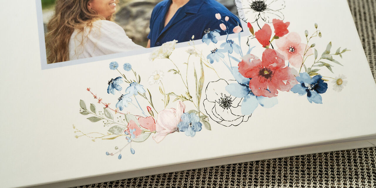 Bližnji posnetek naslovnice knjige gostov s fotografijo mladega para. Elegantna knjiga je opremljena s cvetličnim poročnim motivom in okrasnim premazom na beli podlagi.